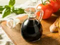 Mały dzbanuszek z ciemnym, gęstym **octem balsamicznym** obok pomidorów, bazylii i grzanek.