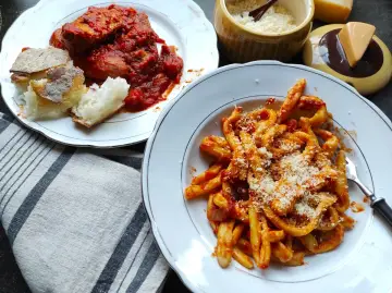 Pyszne maccheroni al ferretto w sosie pomidorowym, posypane parmezanem, obok talerz mięsa w sosie i chleba.