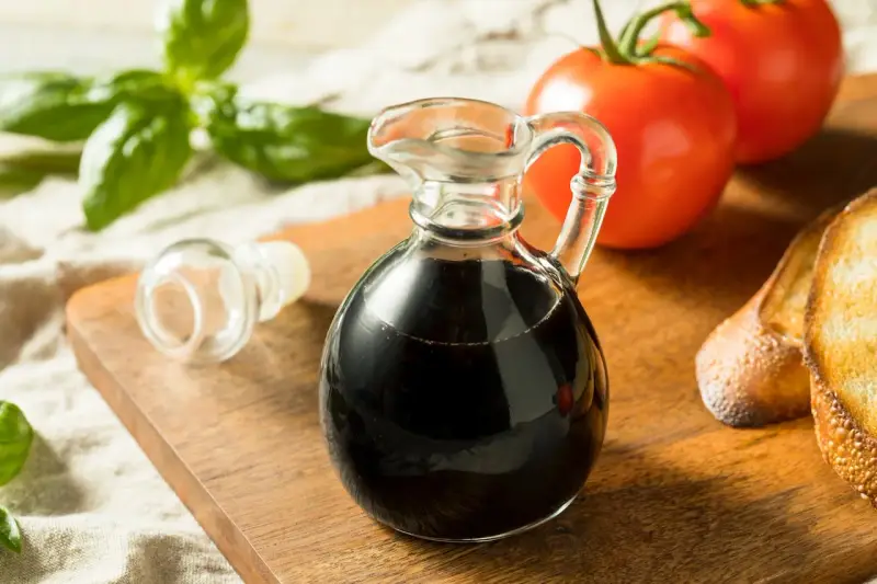 Mały dzbanuszek z ciemnym, gęstym **octem balsamicznym** obok pomidorów, bazylii i grzanek.